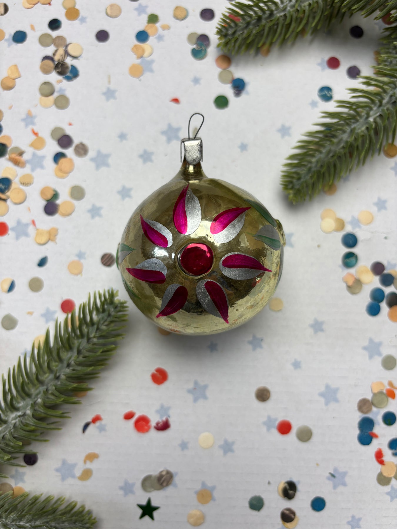 A decorative Christmas ornament with a red gem in the center, surrounded by confetti and pine branches.