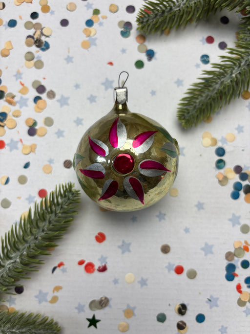 A decorative Christmas ornament with a red gem in the center, surrounded by confetti and pine branches.