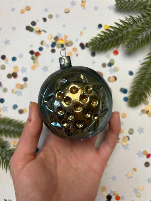 A hand holding a round ornament with a sun-like design, surrounded by colorful confetti and a sprig of greenery.