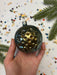 A hand holding a round ornament with a sun-like design, surrounded by colorful confetti and a sprig of greenery.