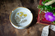 a white ceramic ashtray with a floral design, placed on a wooden surface next to a red book with a floral pattern and a purple flower.