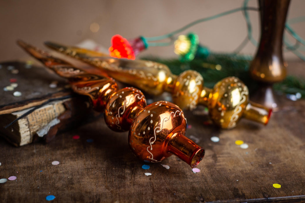 a close-up view of a shiny, golden ornament with a pointed end, resting on a wooden surface. The ornament appears to be a decorative piece, possibly a Christmas ornament or a decorative item. The background is blurred, but it seems to be a festive setting with colorful lights and decorations, suggesting a holiday or celebratory atmosphere.