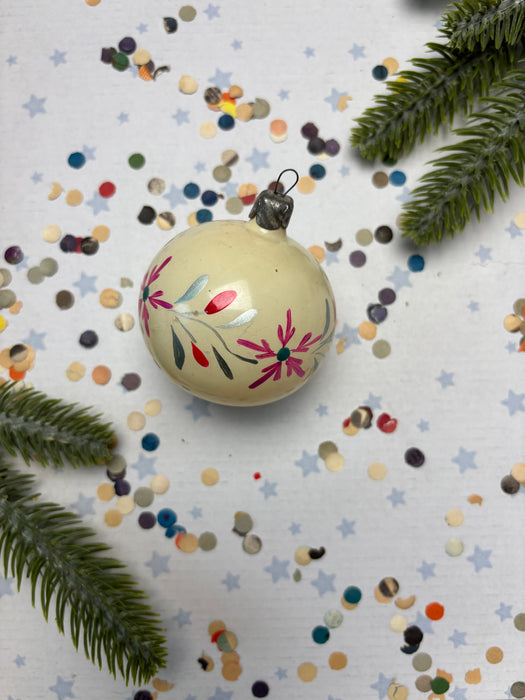 A decorative Christmas ornament with a floral design sits on a table covered in colorful confetti and surrounded by pine branches.