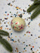 A decorative Christmas ornament with a floral design sits on a table covered in colorful confetti and surrounded by pine branches.