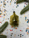 a golden ornament with a black clasp, surrounded by a scattering of colorful confetti and green pine branches.