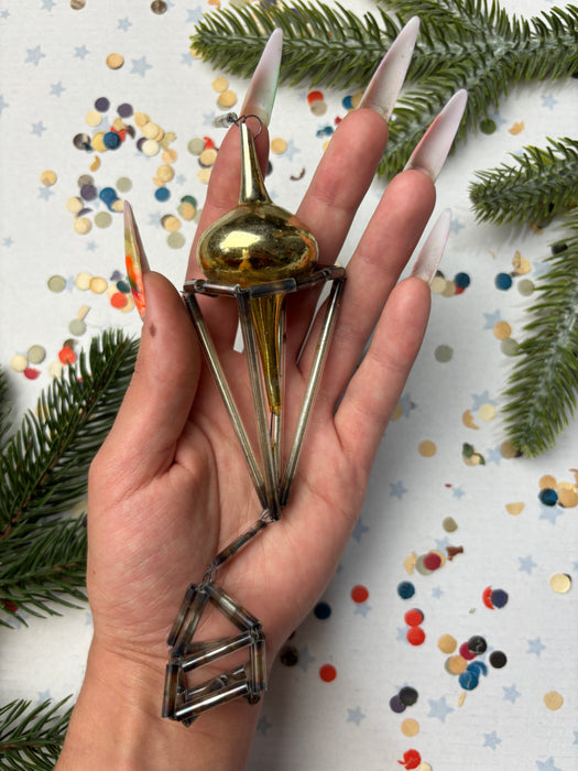 A hand holding a metallic object with a pointed end, surrounded by pine branches and confetti on a white background.