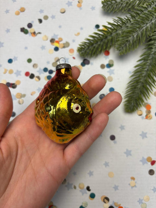 A hand holding a small, shiny, golden ornament resembling a fish against a background of colorful confetti and a pine tree branch.
