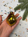 A hand holding a small, shiny, golden ornament resembling a fish against a background of colorful confetti and a pine tree branch.