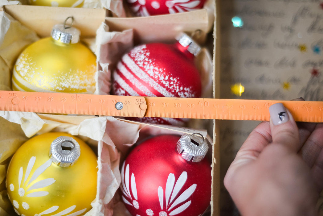 A hand is holding a ruler, measuring the size of a box filled with various ornaments, including yellow and red ones with white designs.