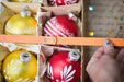 A hand is holding a ruler, measuring the size of a box filled with various ornaments, including yellow and red ones with white designs.