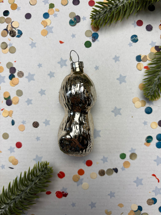 A shiny silver ornament is placed on a white background, surrounded by colorful confetti and green pine branches.