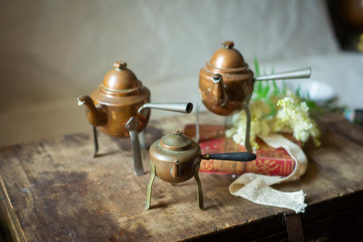 a wooden table with three small copper teapots and a book, along with some white flowers.