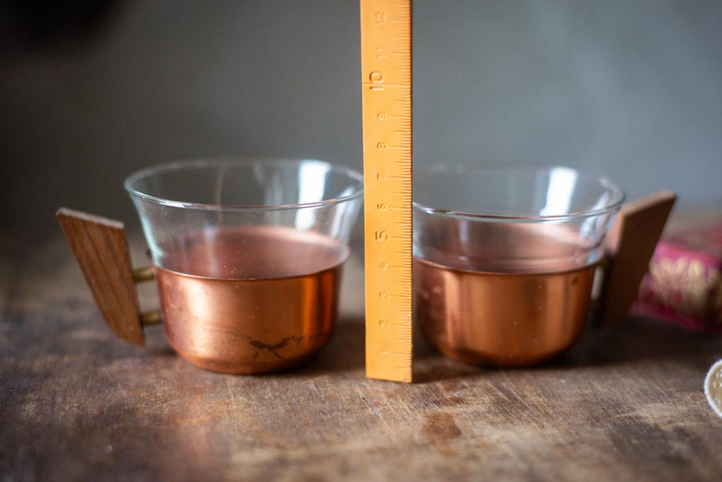 Two copper-colored cups with wooden handles are placed next to a yellow ruler on a wooden surface.