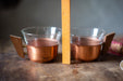 Two copper-colored cups with wooden handles are placed next to a yellow ruler on a wooden surface.
