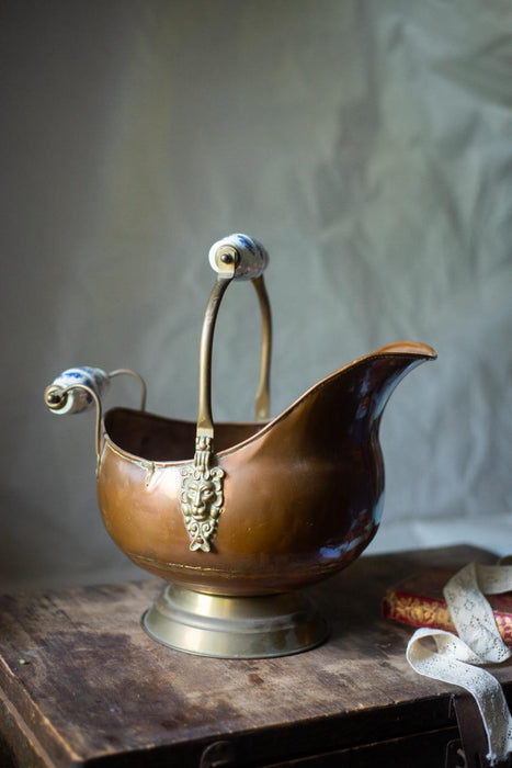 An antique brass kettle with a decorative handle and spout sits on a wooden surface, with a white cloth visible in the background.