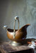 An antique brass kettle with a decorative handle and spout sits on a wooden surface, with a white cloth visible in the background.