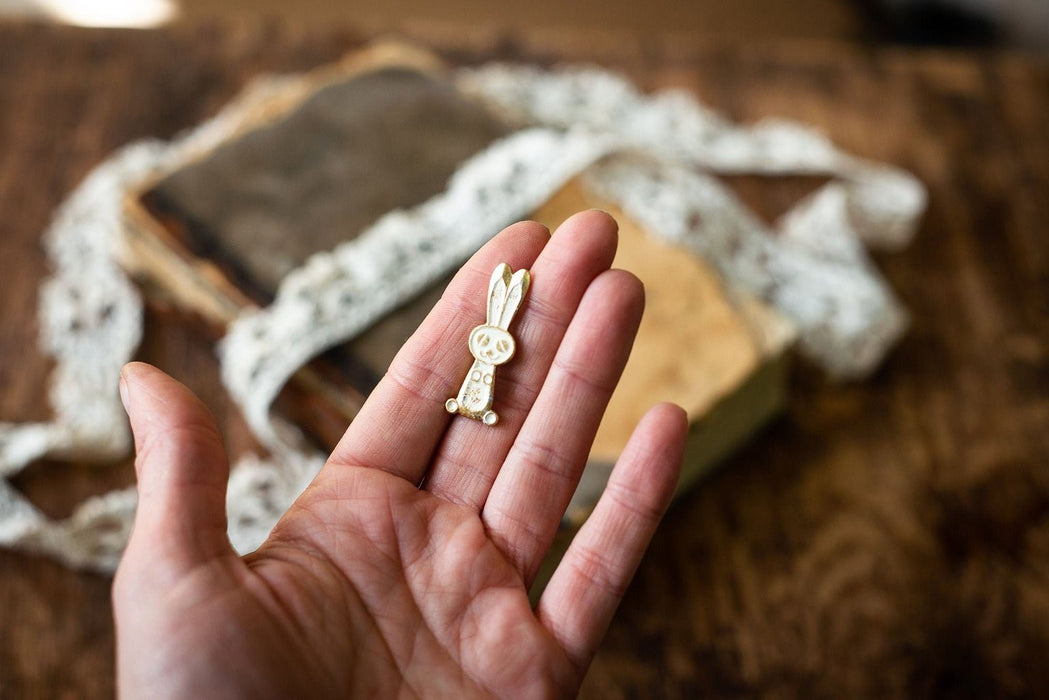 A hand holding a small white rabbit charm against a blurred background of books and lace.