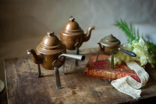 three antique copper teapots placed on a wooden surface, with a red book and a white lace doily in the background.
