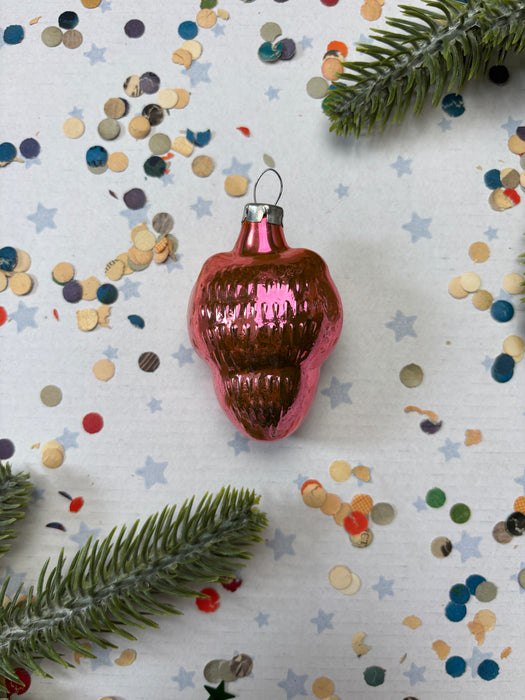 A pink ornament with a shiny surface sits on a white background, surrounded by scattered confetti and a sprig of greenery.