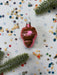 A pink ornament with a shiny surface sits on a white background, surrounded by scattered confetti and a sprig of greenery.