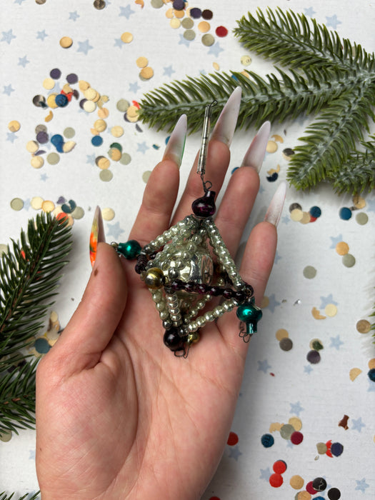A hand holding a beaded necklace with a metallic pendant, surrounded by pine branches and confetti.