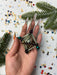 A hand holding a beaded necklace with a metallic pendant, surrounded by pine branches and confetti.