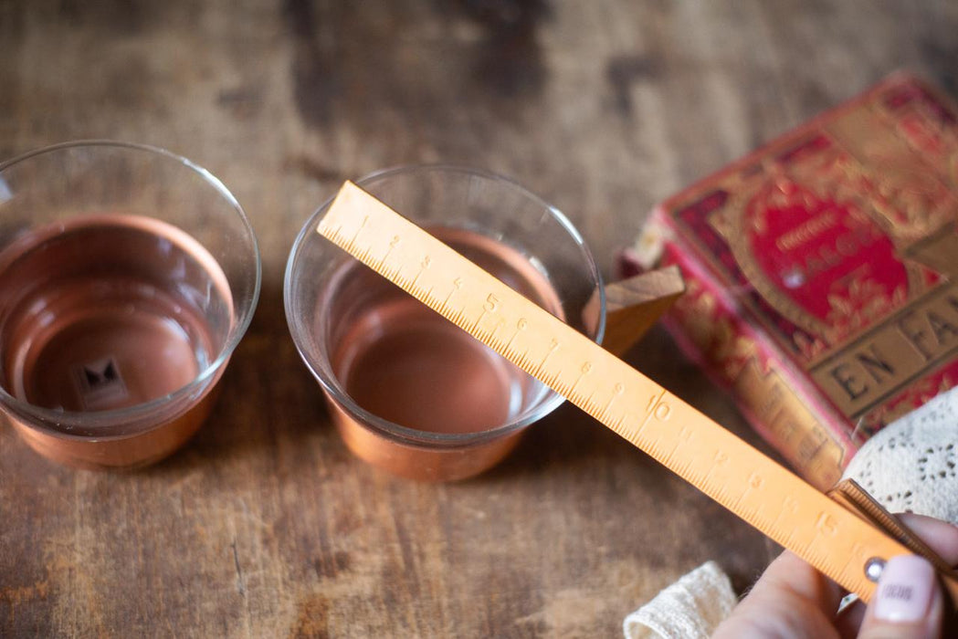 two copper-colored bowls filled with a reddish-brown liquid, placed on a wooden surface. A wooden ruler is being used to measure the volume of the liquid in the bowls.
