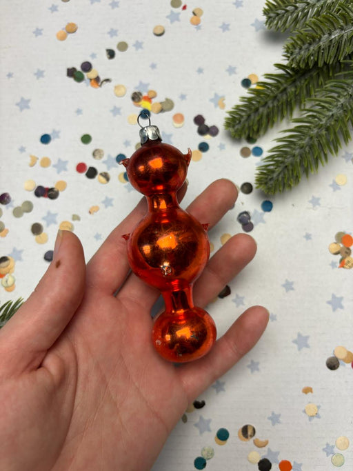 A hand holding a red glass ornament with a silver finial, surrounded by scattered confetti and a sprig of greenery.