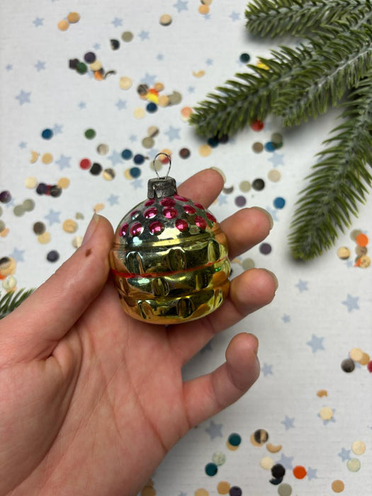 A hand holding a small, colorful ornament with a shiny, metallic appearance, surrounded by scattered confetti and a sprig of greenery.