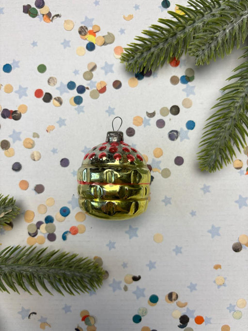 A shiny gold ornament with red and blue accents is surrounded by colorful confetti and pine branches on a white background.