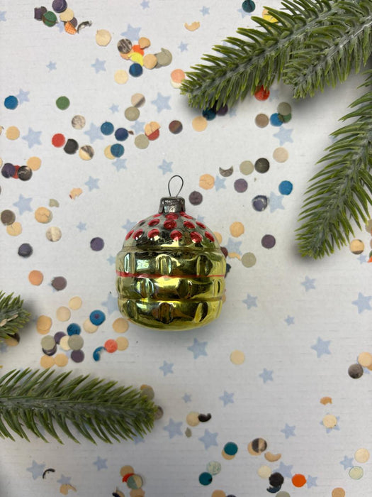 A shiny gold ornament with red and blue accents is surrounded by colorful confetti and pine branches on a white background.