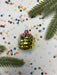 A shiny gold ornament with red and blue accents is surrounded by colorful confetti and pine branches on a white background.