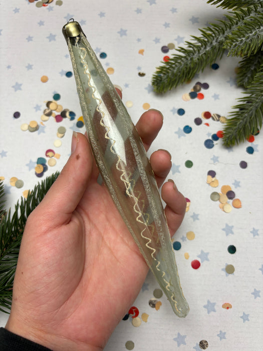 A hand holding a clear glass ornament with a wavy design, surrounded by colorful confetti and pine branches.