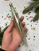 A hand holding a clear glass ornament with a wavy design, surrounded by colorful confetti and pine branches.
