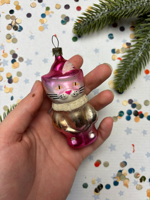 A hand holding a pink and gold ornament with a cat face, surrounded by confetti and a pine branch.
