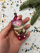 A hand holding a pink and gold ornament with a cat face, surrounded by confetti and a pine branch.