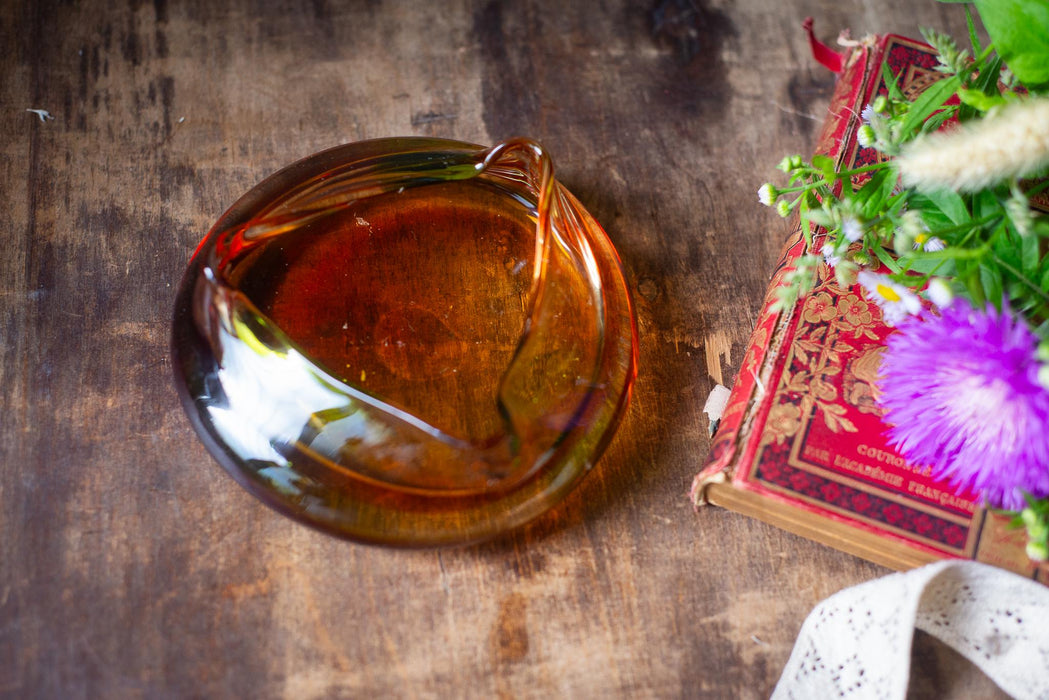 a glass ashtray with a piece of paper inside, placed on a wooden surface next to a book with a purple flower.
