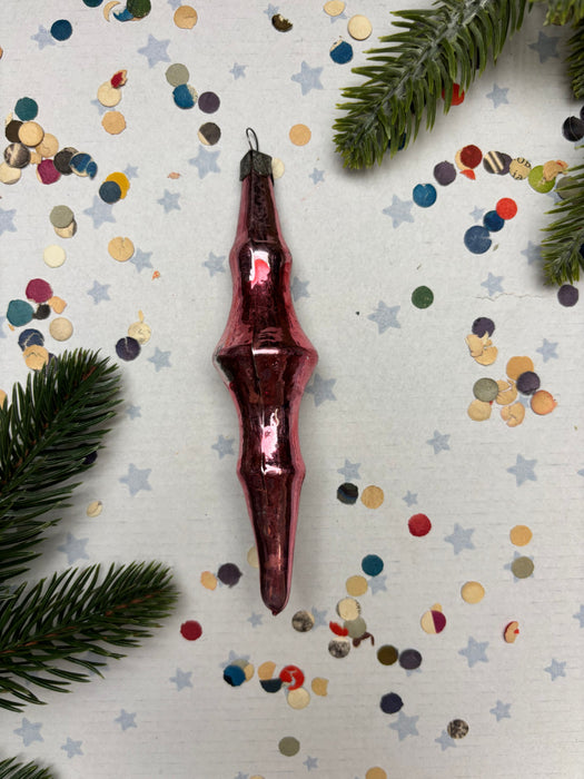 A red glass ornament with a pointed end is surrounded by colorful confetti on a white background, with a pine branch visible on the left side.