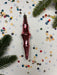 A red glass ornament with a pointed end is surrounded by colorful confetti on a white background, with a pine branch visible on the left side.