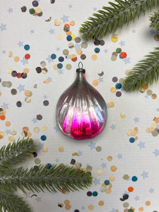 A pink glass ornament with a reflective surface is surrounded by colorful confetti and green pine branches against a white background.