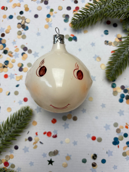 A white ornament with a red smiley face is surrounded by colorful confetti and pine branches on a white background.