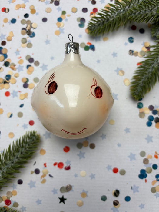 A white ornament with a red smiley face is surrounded by colorful confetti and pine branches on a white background.