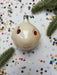 A white ornament with a red smiley face is surrounded by colorful confetti and pine branches on a white background.
