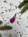A pink ornament is surrounded by colorful confetti and green pine branches on a white background.