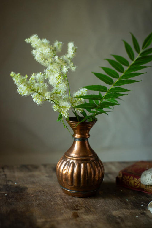 A copper vase holding white flowers and green leaves sits on a wooden surface, with a red and gold patterned cloth visible in the background.