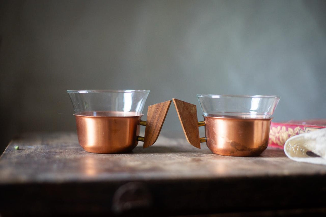 Two copper-colored mugs with wooden handles are placed on a wooden surface, with a blurred background.