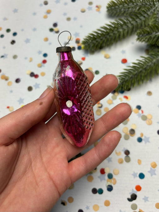 A hand holding a pink glass ornament with a metallic texture, surrounded by colorful confetti on a festive background.