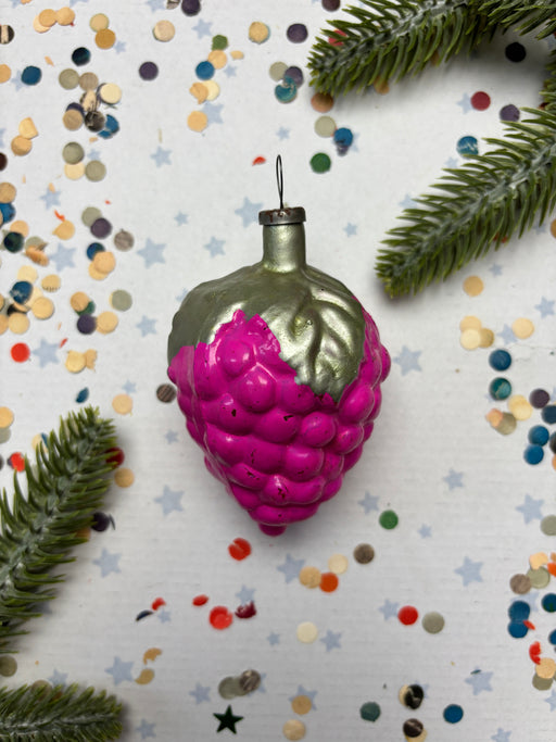 A pink grape ornament with a green stem is surrounded by confetti and pine branches on a white background.