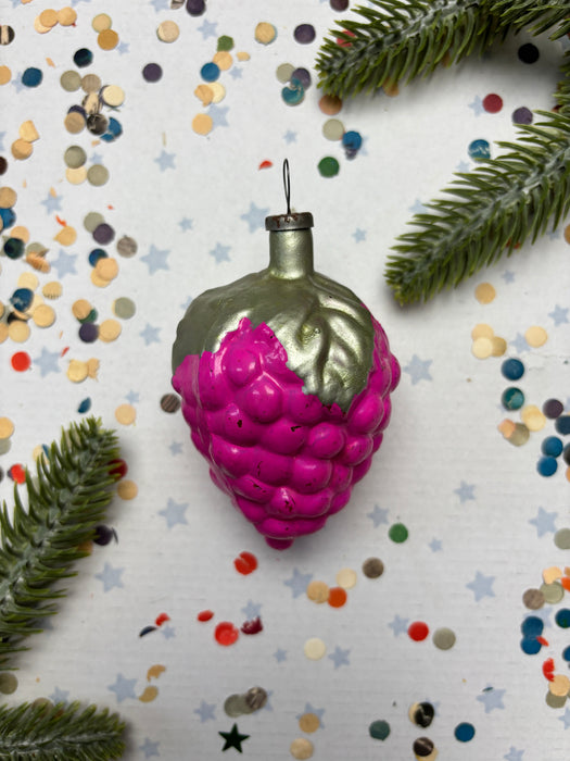 A pink grape ornament with a green stem is surrounded by confetti and pine branches on a white background.