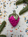 A pink grape ornament with a green stem is surrounded by confetti and pine branches on a white background.